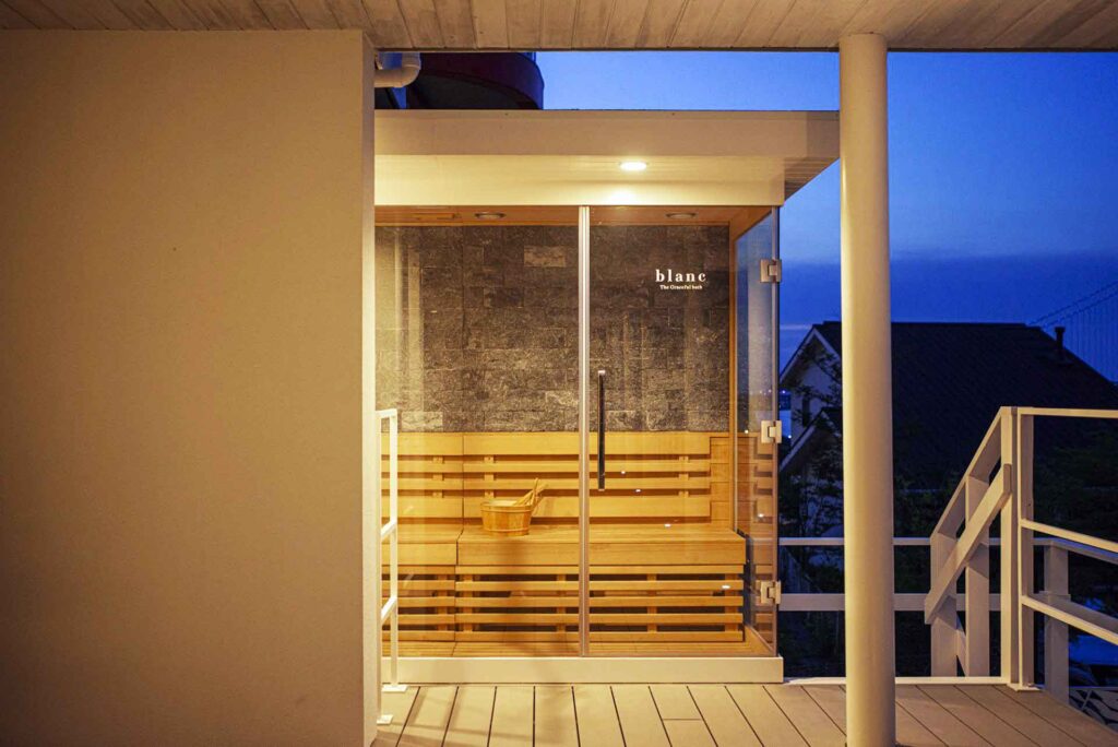 夕暮れ時の屋外に設置された、モダンなガラス張りのサウナルームです。サウナの内部は木製で、ガラスのドアには「blanc The Grand’s Deck」という文字が見えます。サウナルームは、ヴィラの建物とつながるウッドデッキの上にあります。
