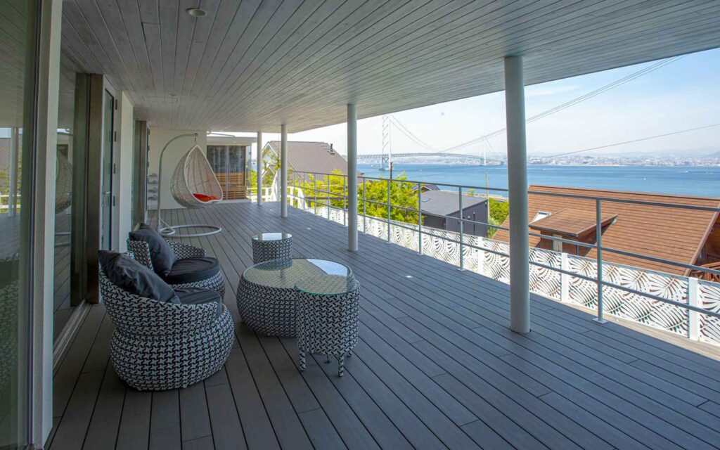 日の光が差し込む広々としたテラスです。テラスには、複数のラタン風の椅子とテーブルが置かれており、天井からは吊り下げ式のチェアがぶら下がっています。テラスからは、穏やかな海と、その向こうに架かる明石海峡大橋がはっきりと見えます。