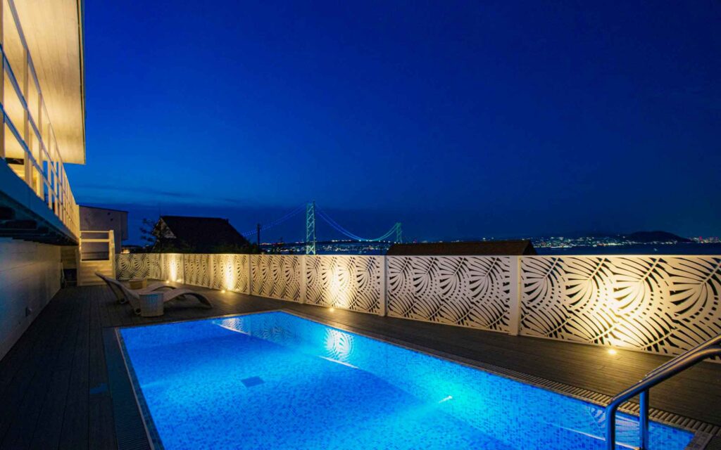 夜の屋外プールエリアです。プールの水面は青くライトアップされ、対岸の街並みと明石海峡大橋のライトアップが輝いています。デッキの端には、葉っぱの模様が彫られた白いフェンスがあり、埋め込み式のライトがフェンスを照らしています。