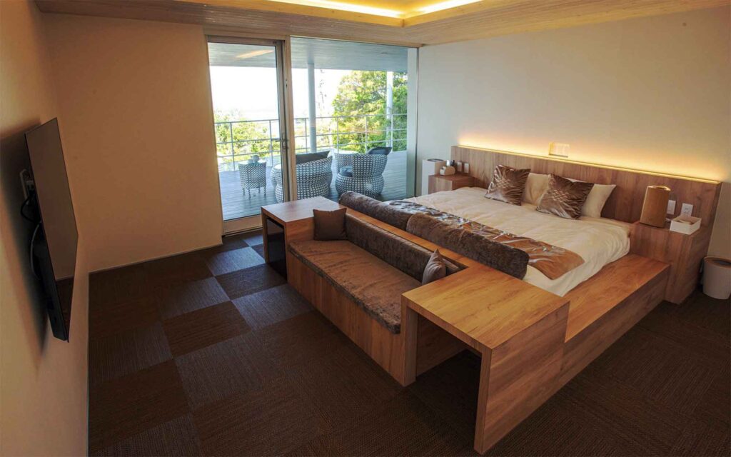 モダンなベッドルームです。大きなキングサイズのベッドは、木製の低いフレームに載っており、床は茶色とベージュの市松模様のタイル張りになっています。ベッドの足元には、同じ木材で作られたベンチと、2つの小さなテーブルが一体になったユニークなデザインの家具が置かれています。大きなガラスの引き戸からは、屋外テラスと海が見えます。