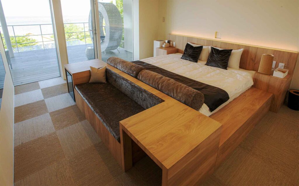 モダンなベッドルームです。木製のプラットフォームベッドは白い寝具と黒いアクセントの枕で飾られています。ベッドの足元には、茶色のクッション付きベンチと木製のテーブルが組み合わされた家具が置かれています。部屋からは大きなガラスの引き戸を通して、屋外テラスと海が眺められます。テラスにはラタン製の吊り下げ式のチェアが見えます。