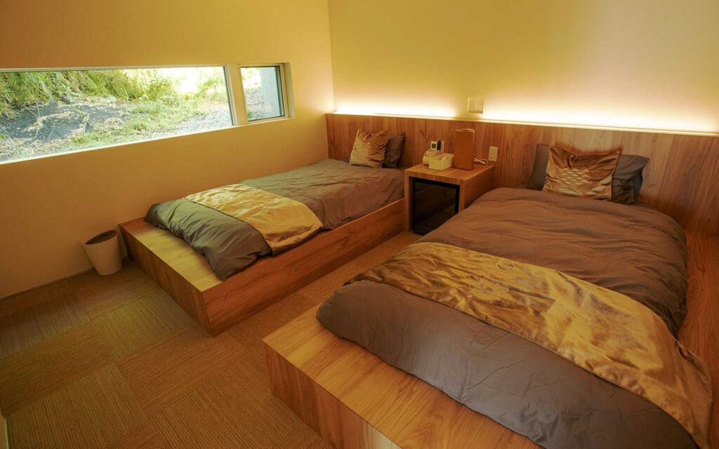 モダンなツインベッドルームです。木製の低いベッドが2つ並んでおり、グレーの掛け布団と金色のスローケットがかけられています。ベッドの間には小さなサイドテーブルが置かれ、ヘッドボードの後ろには間接照明が施されています。