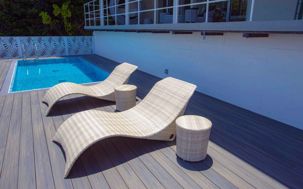 プールの横にあるウッドデッキの写真です。2つの白いラタン製のビーチチェアと、その間に小さな丸いテーブルが置かれています。奥には、青いモザイクタイルが敷かれた長方形のプールが見えます。