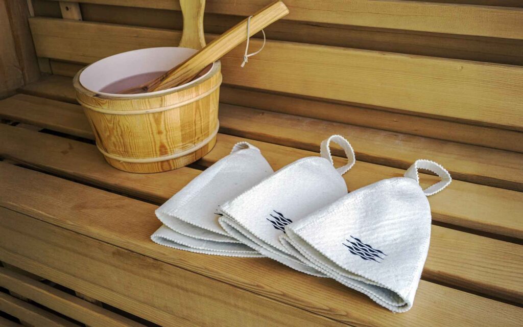 サウナルームの木製ベンチに、サウナハットと木製の桶が置かれています。3つの白いサウナハットには、青い波のような模様が刺繍されており、桶の中には水と柄杓が入っています。