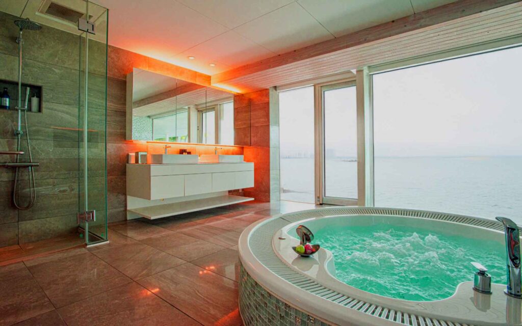 海に面した豪華なバスルームです。手前には、水面が泡立つ大きな円形のジャグジーがあり、大きな窓からは海と対岸の街並みが見えます。左側にはガラス張りのシャワールームと、2つのシンクがある白い洗面台があります。