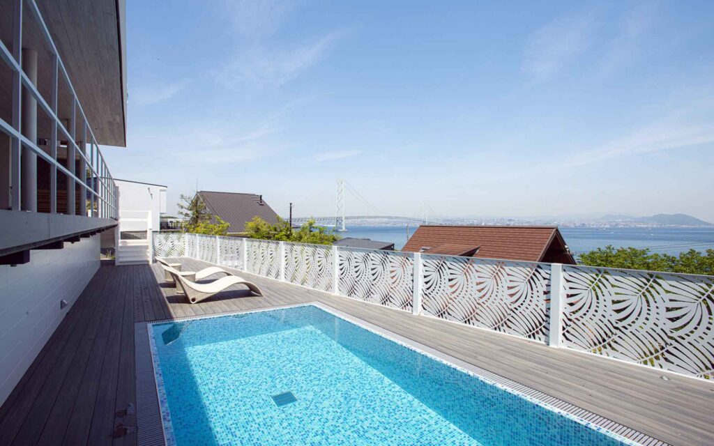 はい、承知いたしました。これらの写真のALTテキストを作成します。
提供された画像のALTテキスト
1. 1F-9.jpg
晴れた日の屋外プールエリアです。ウッドデッキには、青いモザイクタイルが敷かれた長方形のプールがあり、その横に白いビーチチェアが2つ置かれています。テラスの端にある白い手すりには、葉っぱのような模様の切り抜きがあり、その向こうには海と明石海峡大橋が見えます。