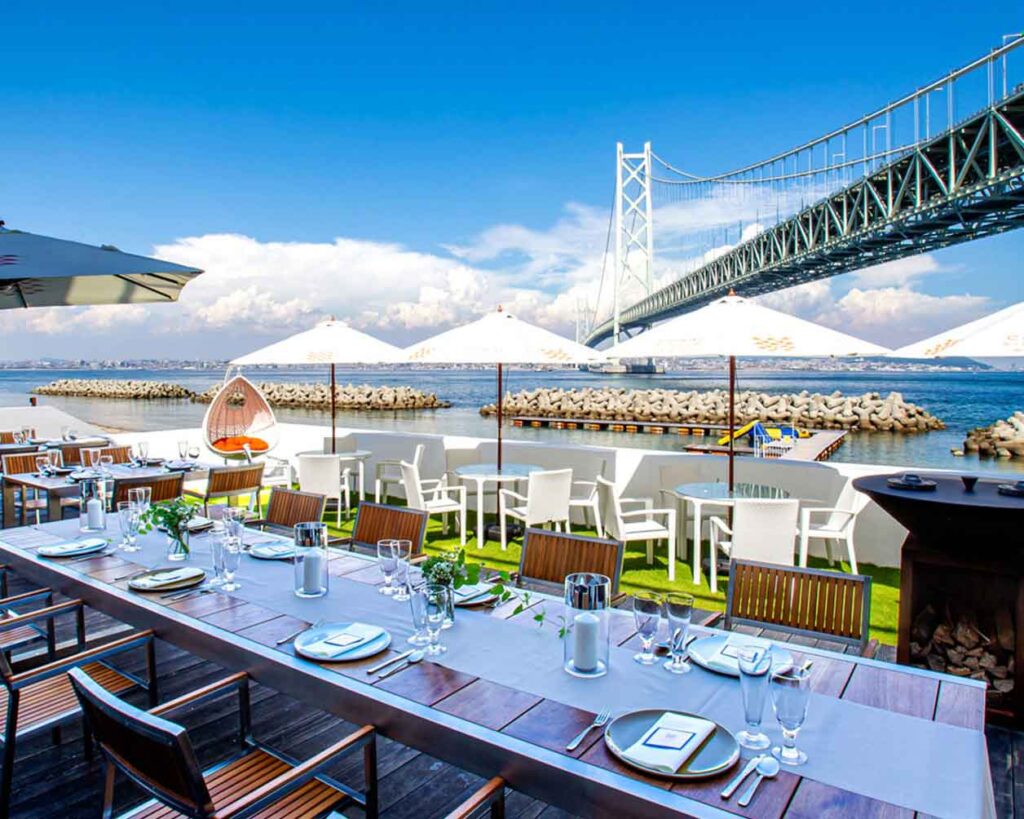 晴れた日の長い屋外ダイニングエリアです。木製のデッキと芝生エリアには、いくつかのテーブルがあり、一部には白いパラソルが立てられています。背景には、青い空といくつかの雲の下に、海と壮大な吊り橋が見えます。