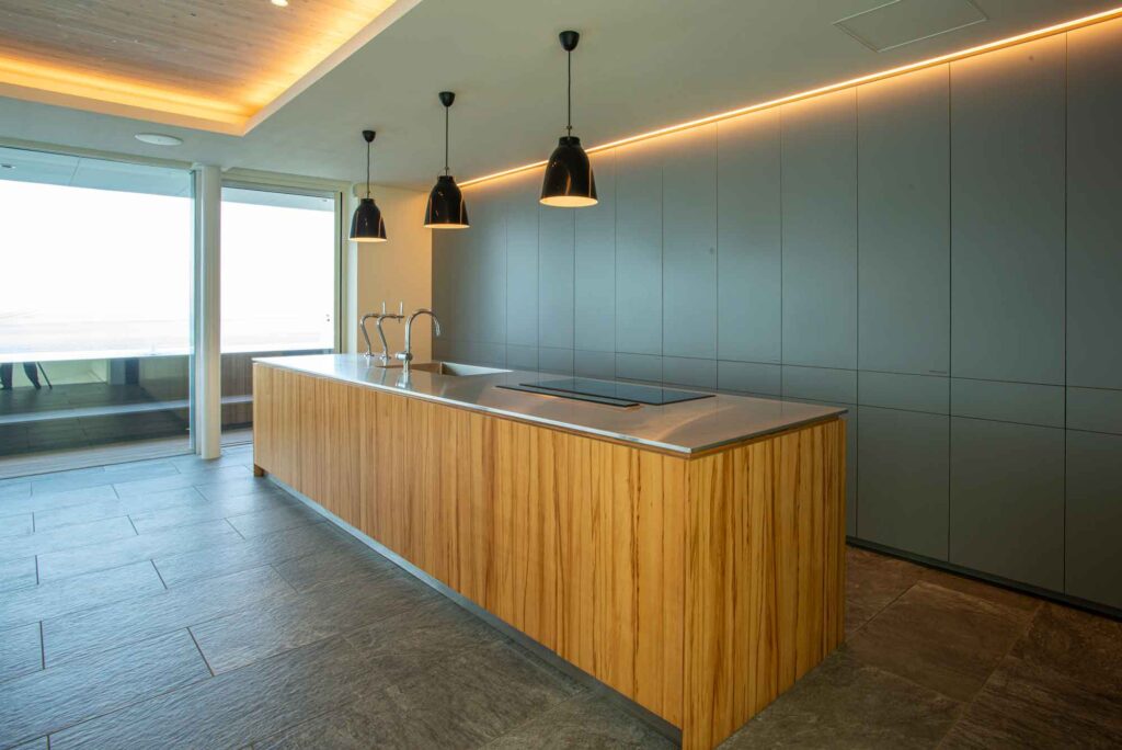 広々とした2階のリビング・ダイニングスペースです。手前にはピンクのクッションが置かれた大きな半円形のソファがあり、中央には円形のガラスのコーヒーテーブルがあります。奥にはダイニングテーブルと椅子、そしてキッチンカウンターが見えます。天井には埋め込み式の照明と木製のパネルが施されています。