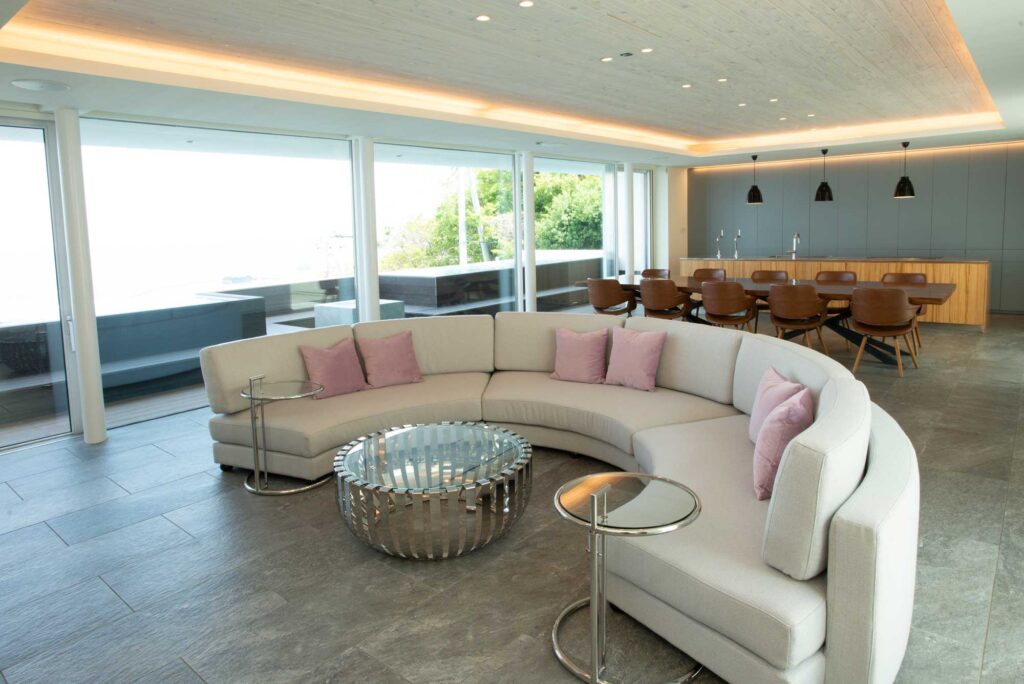 広々とした2階のリビング・ダイニングスペースです。手前にはピンクのクッションが置かれた大きな半円形のソファがあり、中央には円形のガラスのコーヒーテーブルがあります。奥にはダイニングテーブルと椅子、そしてキッチンカウンターが見えます。天井には埋め込み式の照明と木製のパネルが施されています。