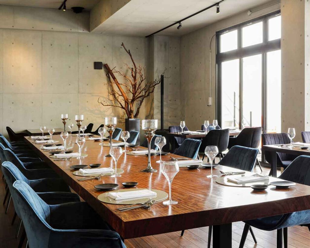 モダンなレストランの店内で、グループ用にセットされた長く大きな木製のダイニングテーブルがあります。部屋はコンクリートの壁で、自然光が差し込む大きな窓があります。濃い青のベルベットの椅子がテーブル沿いに置かれ、テーブルには複数のグラスセットと背の高い燭台のようなセンターピースが飾られています。