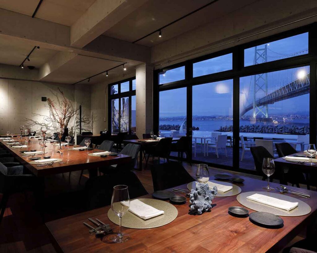 夕暮れ時のモダンなレストランの店内です。長い木製のダイニングテーブルには、皿、グラス、カトラリーがセットされ、その周りには濃い色の椅子が並んでいます。右側の大きな窓からは、夜空にライトアップされた吊り橋と水面の街の明かりが見える素晴らしい景色が広がっています。