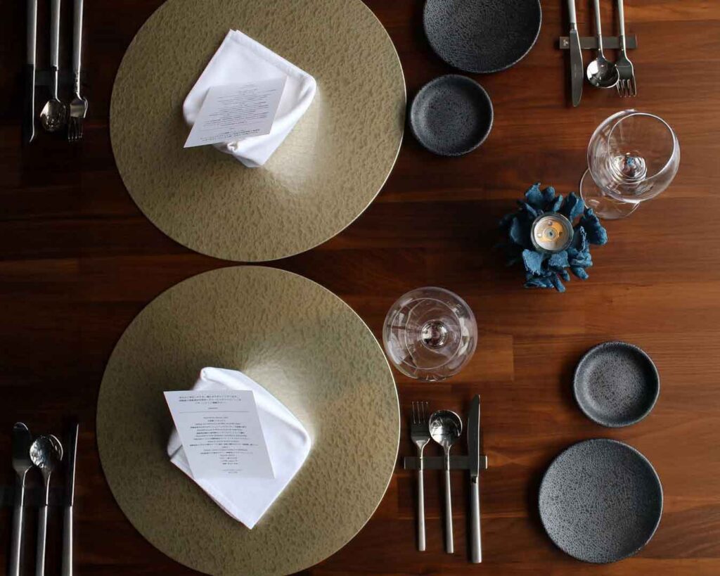 暗い木製のテーブルに置かれた2つのテーブルセッティングを上から撮影した写真です。それぞれのセッティングには、質感のある金色のプレースマット、白い折りたたまれたナプキン、そしてメニューが含まれています。濃い灰色の皿と磨かれたカトラリーセットがプレースマットの周りにきれいに配置されています。2つのセッティングの間には、灯されたキャンドルが入った小さな青い飾り物が置かれています。