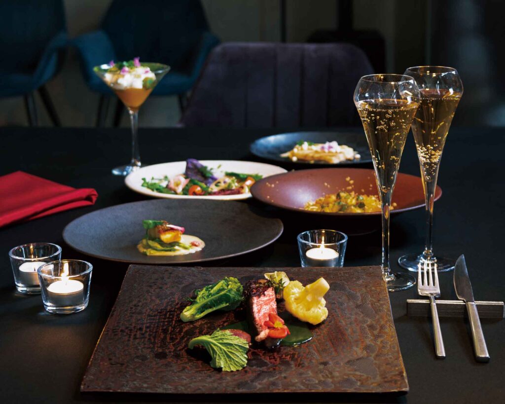 暗いテーブルに美しく盛り付けられた料理のクローズアップです。手前の四角い皿には、焼かれた肉と緑と黄色の野菜が添えられています。その後ろには、他にも数品のアートのように配置された料理、2本のスパークリングワインのフルートグラス、そしていくつかの灯されたティーキャンドルが見えます。