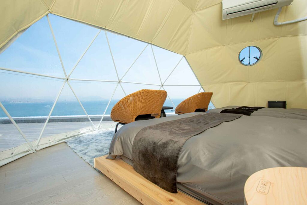 ドーム型テントの寝室内部です。ベッドと2つのラタン製の椅子が、透明な大きな窓の方を向いて配置されています。窓は特徴的な三角形のフレームで区切られており、そこから海と水平線の素晴らしい景色が広がっています。ドームの側面には、船の丸窓のような小さな窓も見えます。