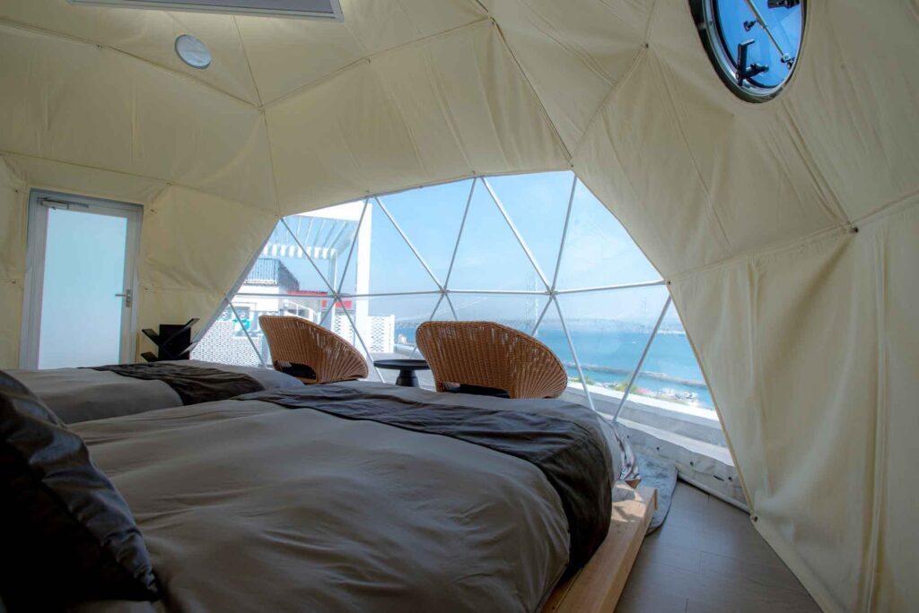 ドーム型テントの寝室内部です。グレーと濃い茶色の掛け布団がかけられた2つのツインベッドがあり、テントの正面にある大きな透明な窓からは、海と明石海峡大橋のパノラマビューが見えます。窓の前には2つのラタン製の椅子が置かれています。