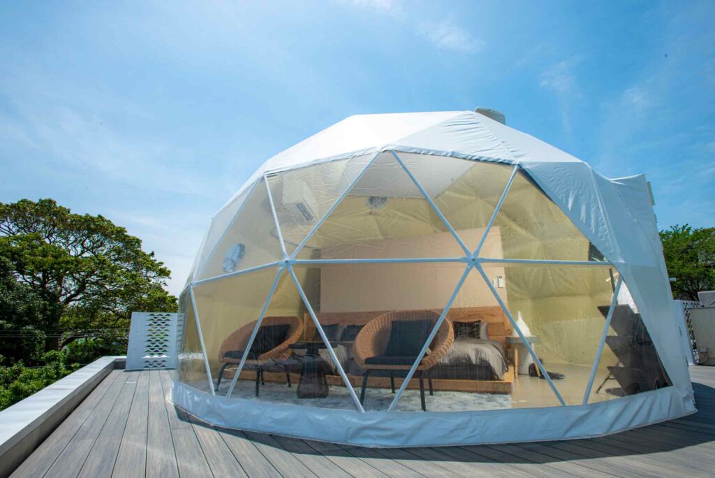 屋上デッキに設置された、ユニークなドーム型テントです。テントの前面は透明で、内部にはベッドと2つのラタン製の椅子が見えます。テントの外側は白色で、ライトウッドの床の上に置かれています。