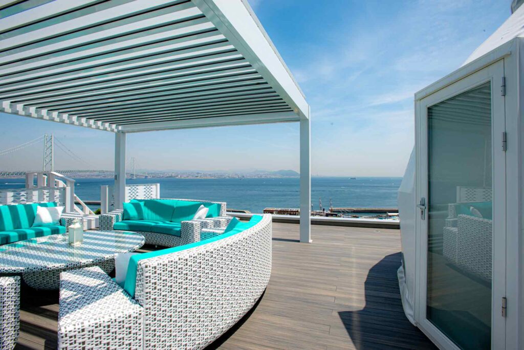 晴れた日の広々とした屋上テラスです。白いラタン製の円形ソファには鮮やかなターコイズ色のクッションが置かれ、それらが中央の丸いテーブルを囲むように配置されています。ソファの上には白い格子状のパーゴラがあり、背景には明石海峡大橋と海が広がる景色が見えます。