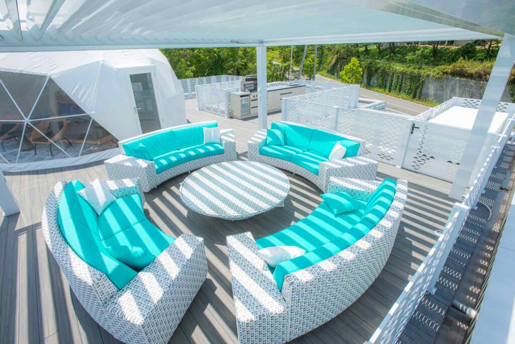屋上テラスを上から撮影した写真です。ターコイズ色のクッションが置かれた白い円形のソファセットが白いパーゴラの下に配置され、その影がクッションとテーブルに縞模様を作っています。テラスには、ドーム型テントと屋外キッチンエリアも見えます。