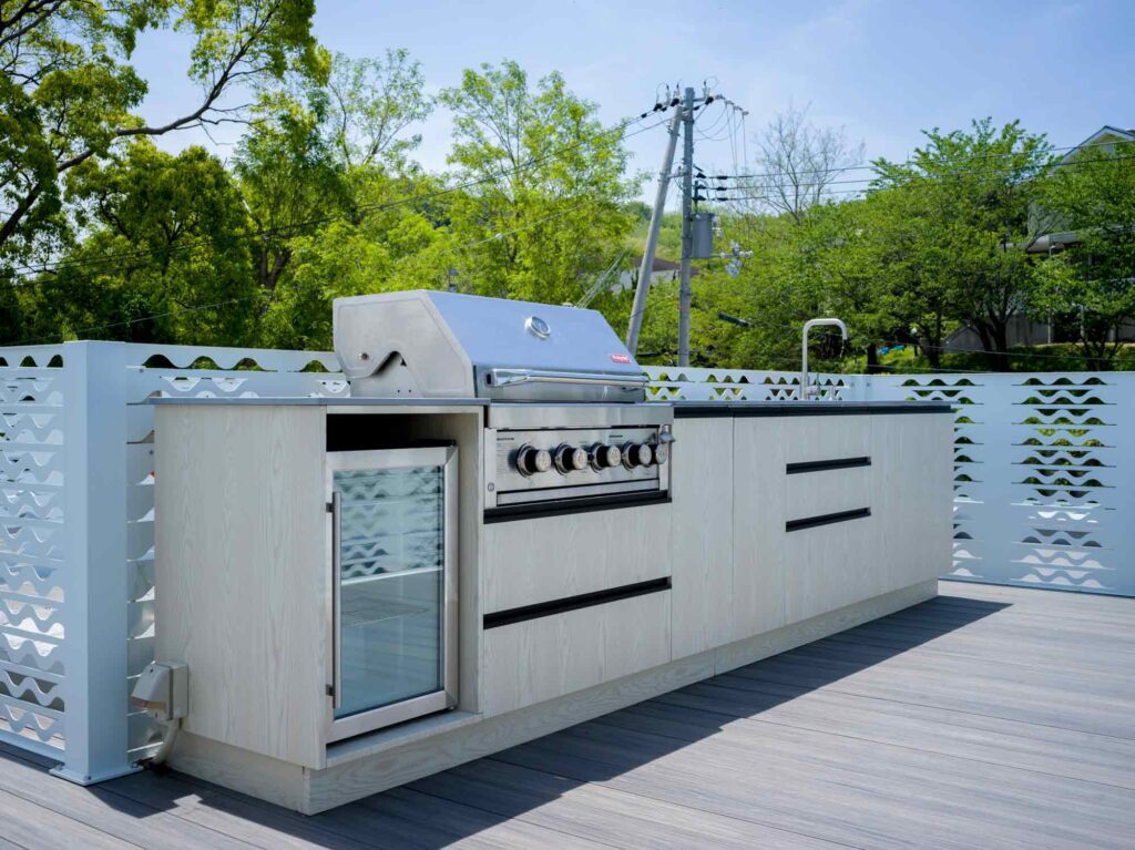 屋上テラスにある屋外キッチンエリアです。明るい木目調のモダンなキッチンカウンターには、グリル、小さな冷蔵庫、そしてシンクが備え付けられています。カウンターの後ろには、波模様が切り抜かれた白い装飾的なフェンスがあります。