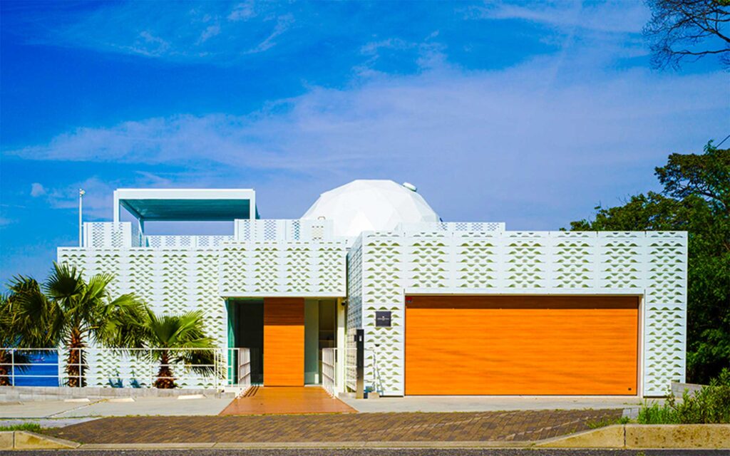 晴れた青空の下、ヴィラのモダンな正面玄関を捉えた外観写真です。白い建物のファサードには、小さな三角形の模様が繰り返されたユニークなデザインのパネルが特徴です。中央には木製の扉があり、右隣には同じ木目調の大きなガレージドアがあります。屋根には白いドーム状の構造物と、上階のルーフトップテラスらしき白いキャノピーが見えます。建物の左側にはヤシの木が植えられています。