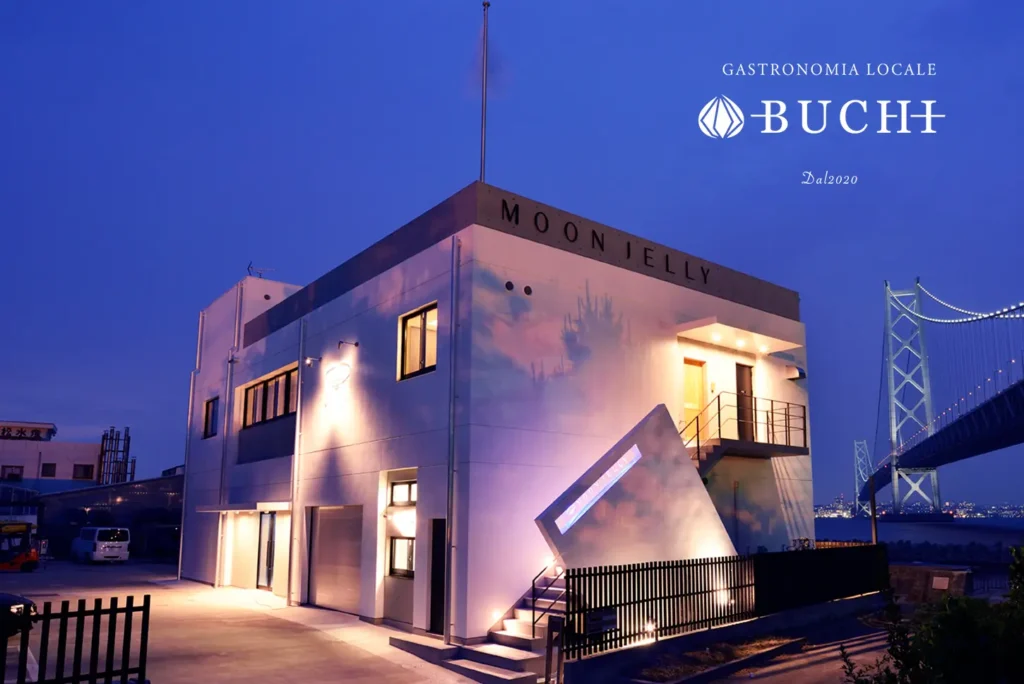 二階建てのモダンな建物が夕暮れ時にライトアップされ、上部には「MOON JELLY」と書かれています。建物のファサードにはカラフルな抽象的な画像が投影されています。右側には、暗い空に照らされた大きな吊り橋が水上に見えます。画像の右上隅には「GASTRONOMIA LOCALE BUCHI Dal.2020」のロゴがあります。