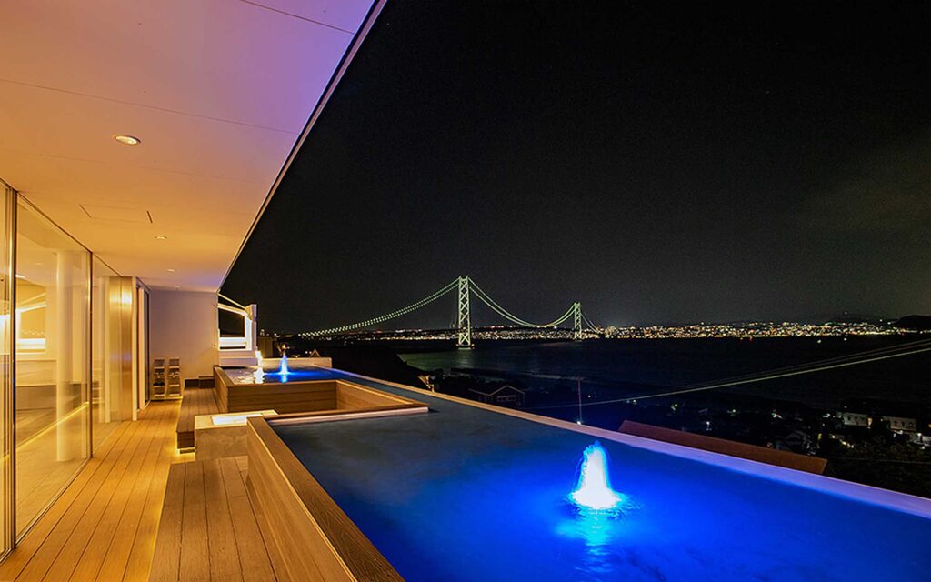 夜空の下、ヴィラの2階にあるインフィニティプールから見た、明石海峡大橋の壮大な夜景です。プールは泳ぐためのものではなく、水面から青く光る噴水が2つ上がっています。プールの奥には、ライトアップされた明石海峡大橋が湾を横切り、その向こうには街の明かりが輝いています。左側には木の床とガラスの壁が並ぶテラスがあり、全体的に落ち着いた豪華な雰囲気を醸し出しています。