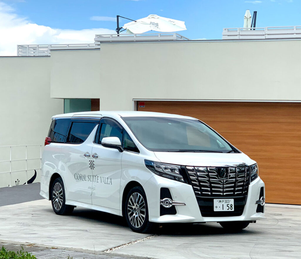 宿泊者専用送迎サービスとして、白いトヨタ・アルファードがモダンな建物の前で停車している様子。運転席側のドアには「CORAL SUITE VILLA」という文字とロゴが書かれている。背景には青空と少し海が見える。